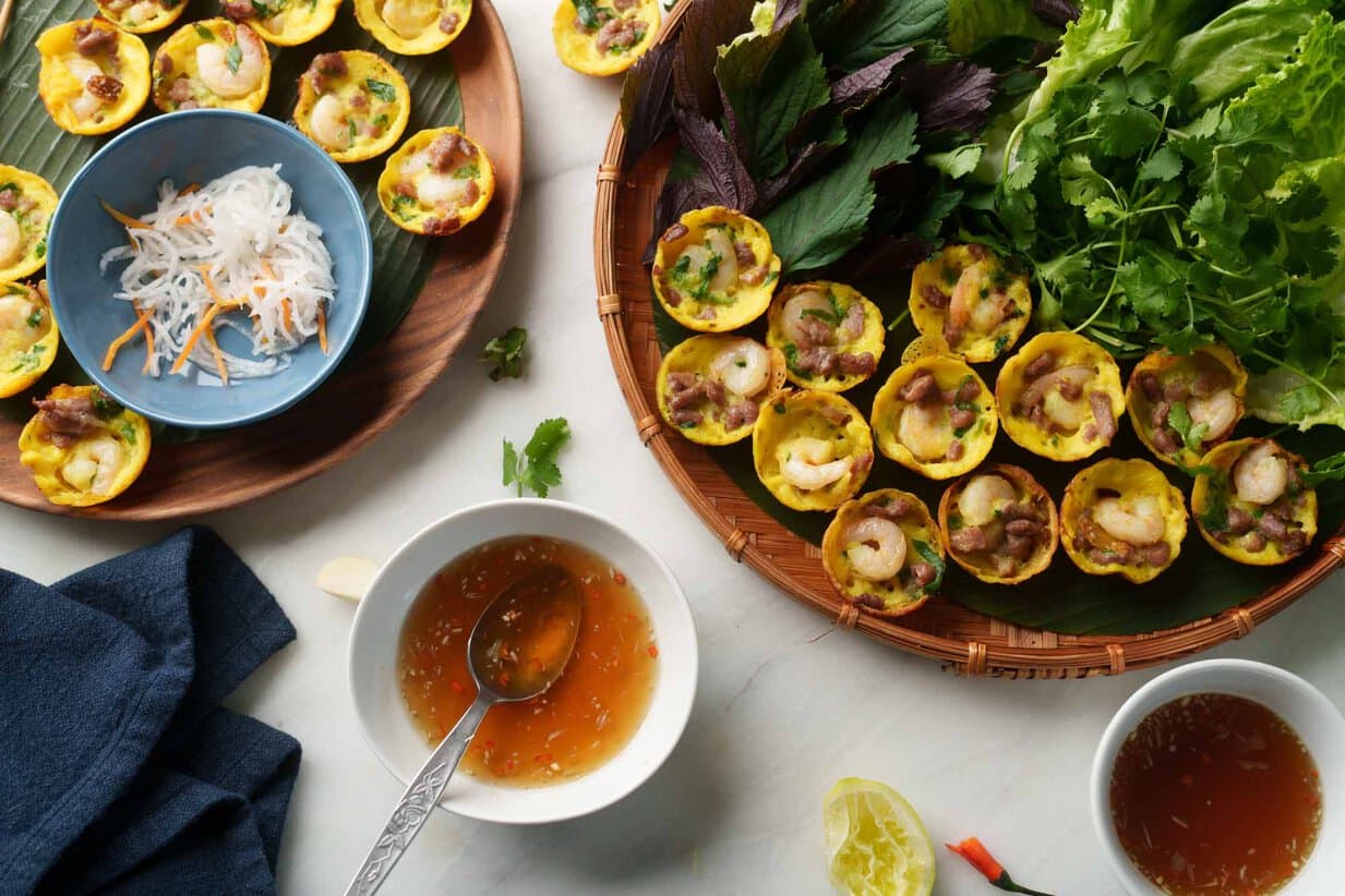 A tray of bánh khọt with dipping sauce and a plate of herbs and lettuce beside it.